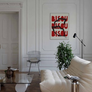 Op de afbeelding: Een witte bank met een wit kussen en een zwarte en zilveren bijzettafel. Een groene plant staat in de hoek van de kamer. Een zwarte en zilveren stoel staat op de achtergrond. Een ingelijst poster met het woord "BISOU" dat drie keer in rode letters wordt herhaald, hangt aan de muur.