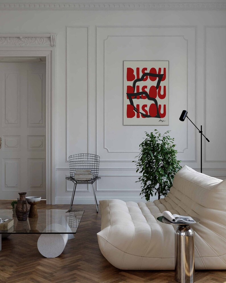 Op de afbeelding: Een witte bank met een wit kussen en een zwarte en zilveren bijzettafel. Een groene plant staat in de hoek van de kamer. Een zwarte en zilveren stoel staat op de achtergrond. Een ingelijst poster met het woord "BISOU" dat drie keer in rode letters wordt herhaald, hangt aan de muur.