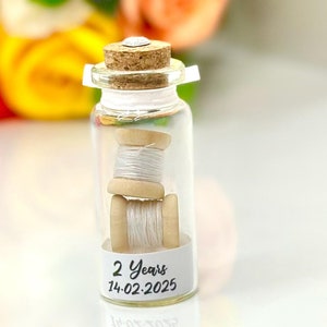 May include: A small glass bottle with a cork stopper contains two spools of white thread. The bottle has a label that reads "2 Years 14.02.2025".