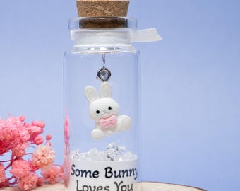 May include: A clear glass bottle with a cork stopper, containing a white bunny figurine with a pink bow. The bottle sits on a wooden slice and has the text "Some Bunny Loves You". Pink flowers are in the background.