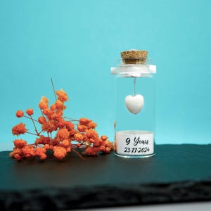 May include: A small glass bottle with a cork stopper contains a white heart charm hanging from a silver chain. The bottle has a label that reads "9 Years 23.11.2024".