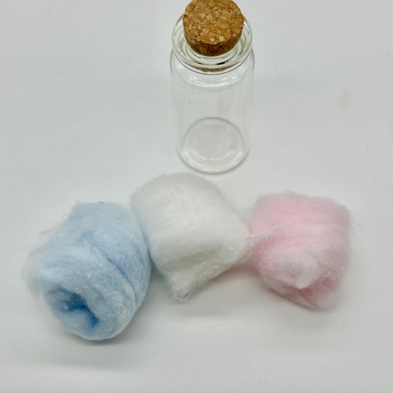 Op de afbeelding: Drie bollen vezel, &eacute;&eacute;n blauwe, &eacute;&eacute;n witte en &eacute;&eacute;n roze, naast een heldere glazen fles met een kurken stop.
