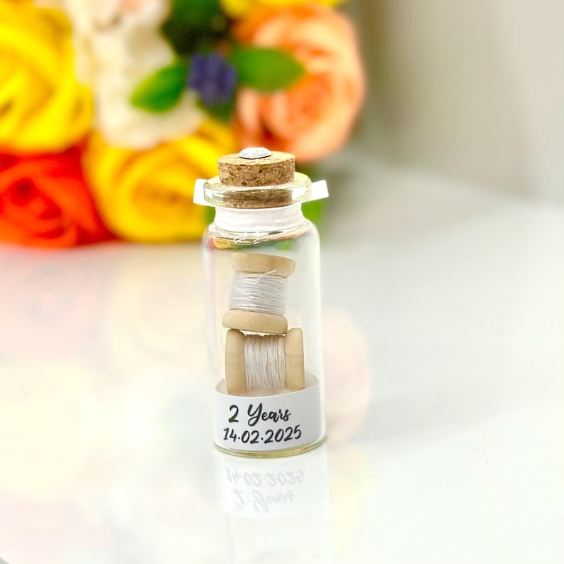 May include: A small glass bottle with a cork stopper contains two spools of white thread. The bottle has a label that reads "2 Years 14.02.2025".