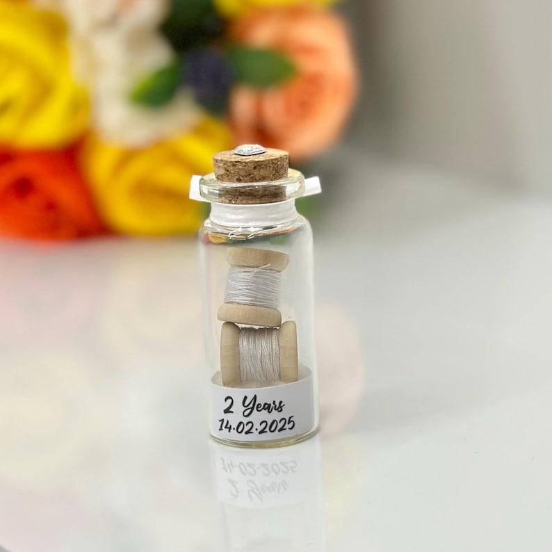 May include: A small glass bottle with a cork top contains two spools of white thread. The bottle has a label that reads "2 Years 14-02-2025".