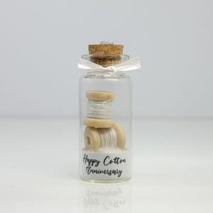 May include: A small glass jar with a cork lid and a pink ribbon tied around the top. Inside the jar are two spools of white thread wrapped around wooden spools. The jar has a white label with the text "Happy Cotton Anniversary".