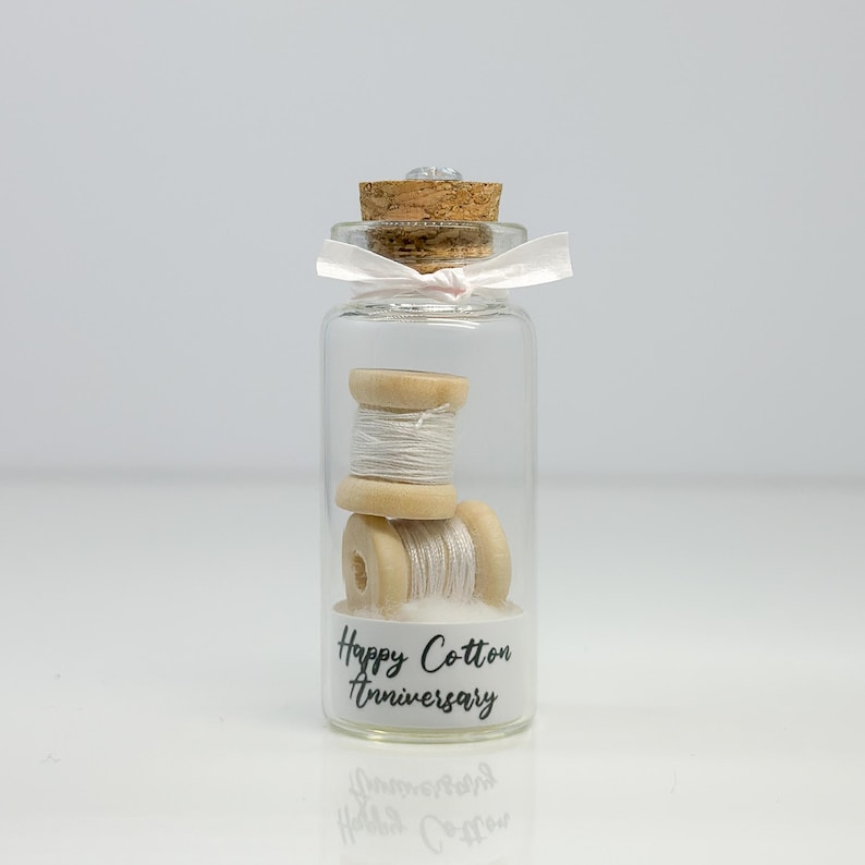 May include: A small glass jar with a cork lid and a pink ribbon tied around the top. Inside the jar are two spools of white thread wrapped around wooden spools. The jar has a white label with the text "Happy Cotton Anniversary".