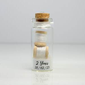 May include: A glass jar with a cork lid contains two spools of white thread. The jar has a label that reads "2 Years 28/02/23".