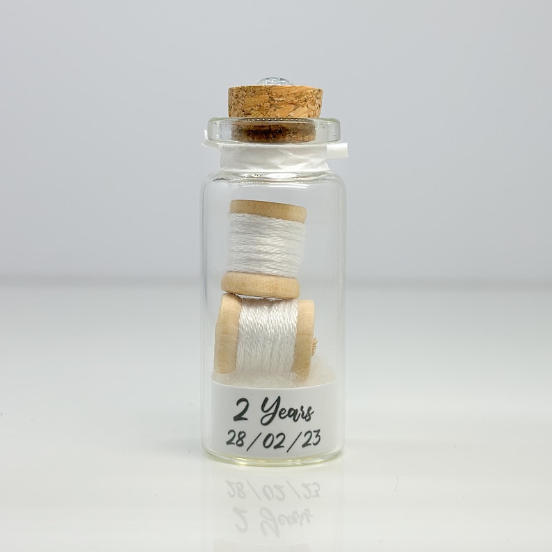 May include: A glass jar with a cork lid contains two spools of white thread. The jar has a label that reads "2 Years 28/02/23".