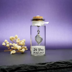 May include: A clear glass bottle with a cork stopper, containing a silver pendant necklace with a white stone and clear crystals. The bottle has a label that reads "24 Years 14.02.2025". A small bouquet of dried flowers sits to the left.