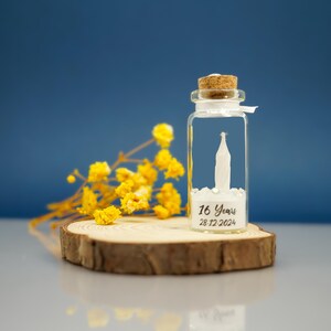 May include: A small glass bottle with a cork stopper, filled with white glitter and a white paper figure. The bottle is sitting on a wooden slice and has a label that reads "16 Years 28.12.2024".