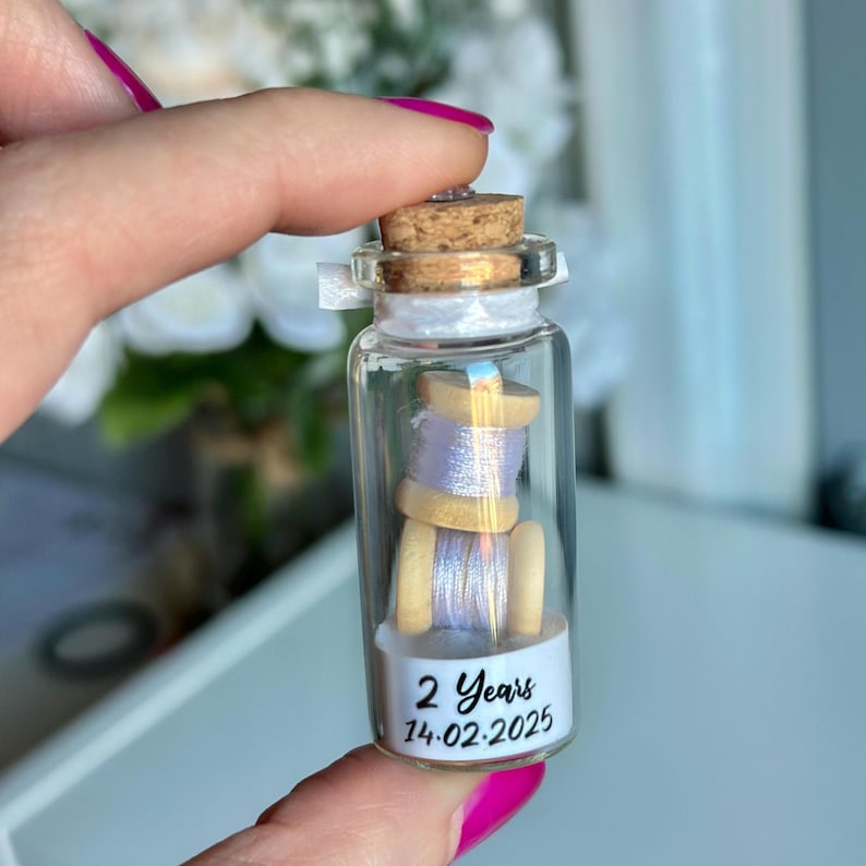 May include: A small glass jar with a cork lid containing two spools of lavender thread. The jar has a white label with the text "2 Years 14.02.2025".