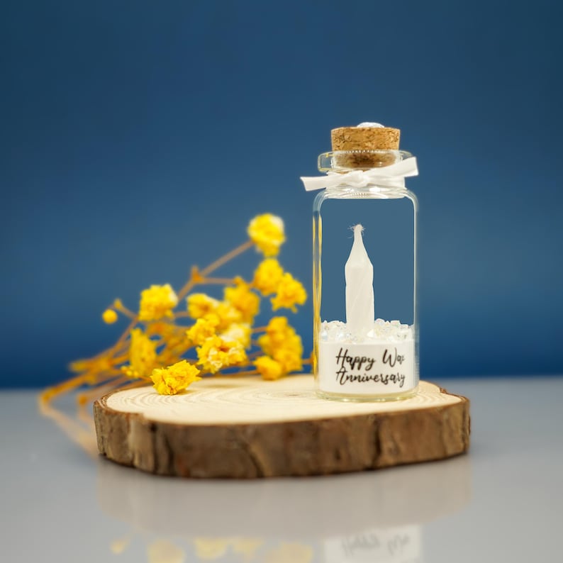 May include: A small glass bottle with a cork stopper and a white ribbon tied around the neck. The bottle contains white glitter and a small white paper figure. The bottle is sitting on a wooden slice and has a label that reads "Happy Wedding Anniversary".