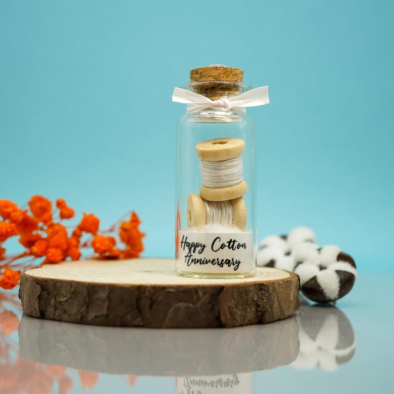 May include: A small glass jar with a cork lid and a white ribbon tied around the top. Inside the jar is a spool of white cotton thread and a small piece of cotton. The jar has a white label with the text "Happy Cotton Anniversary".