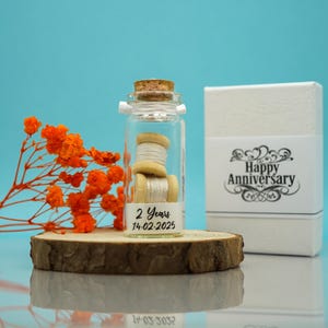 May include: A small glass jar with a cork lid filled with two spools of white thread. The jar has a label that reads "2 Years 14-02-2025". The jar is sitting on a wooden slice. A white box with the text "Happy Anniversary" is in the background.