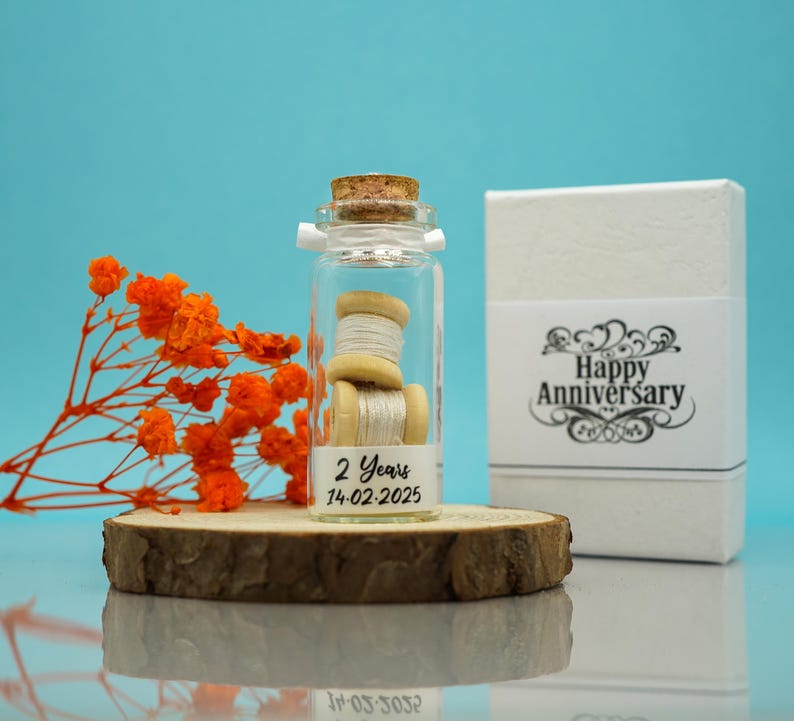 May include: A small glass jar with a cork lid filled with two spools of white thread. The jar has a label that reads "2 Years 14-02-2025". The jar is sitting on a wooden slice. A white box with the text "Happy Anniversary" is in the background.