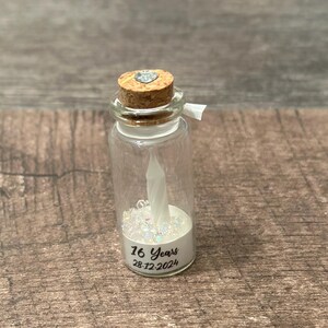 May include: A small glass jar with a cork top and a white heart charm. The jar is filled with white glitter and a white candle. The jar has a label that reads "16 Years 28.12.2024".