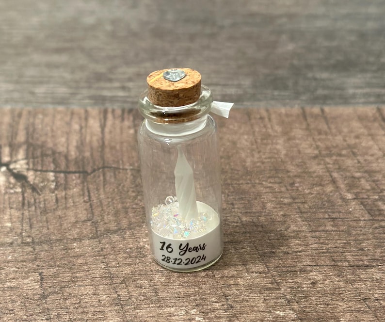 May include: A small glass jar with a cork top and a white heart charm. The jar is filled with white glitter and a white candle. The jar has a label that reads "16 Years 28.12.2024".