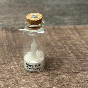 May include: A small glass bottle with a cork top filled with white glitter and a white candle. The bottle has a label that says "Happy Wax Anniversary".