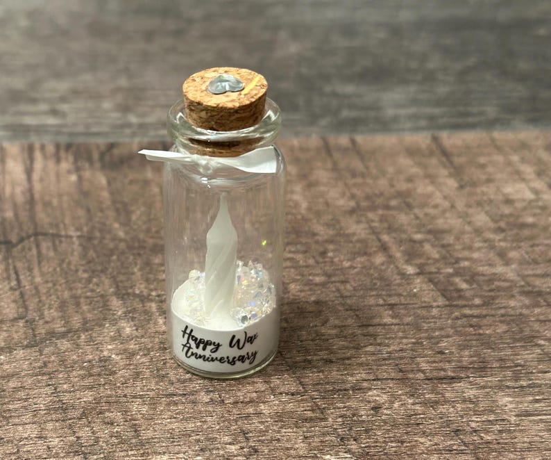 May include: A small glass bottle with a cork top filled with white glitter and a white candle. The bottle has a label that says "Happy Wax Anniversary".