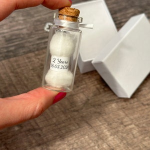 Op de afbeelding: Een klein glazen flesje met een kurkstop en een wit lint. Het flesje bevat twee witte wattenbolletjes en een label met de tekst "2 Years 18.03.2024".
