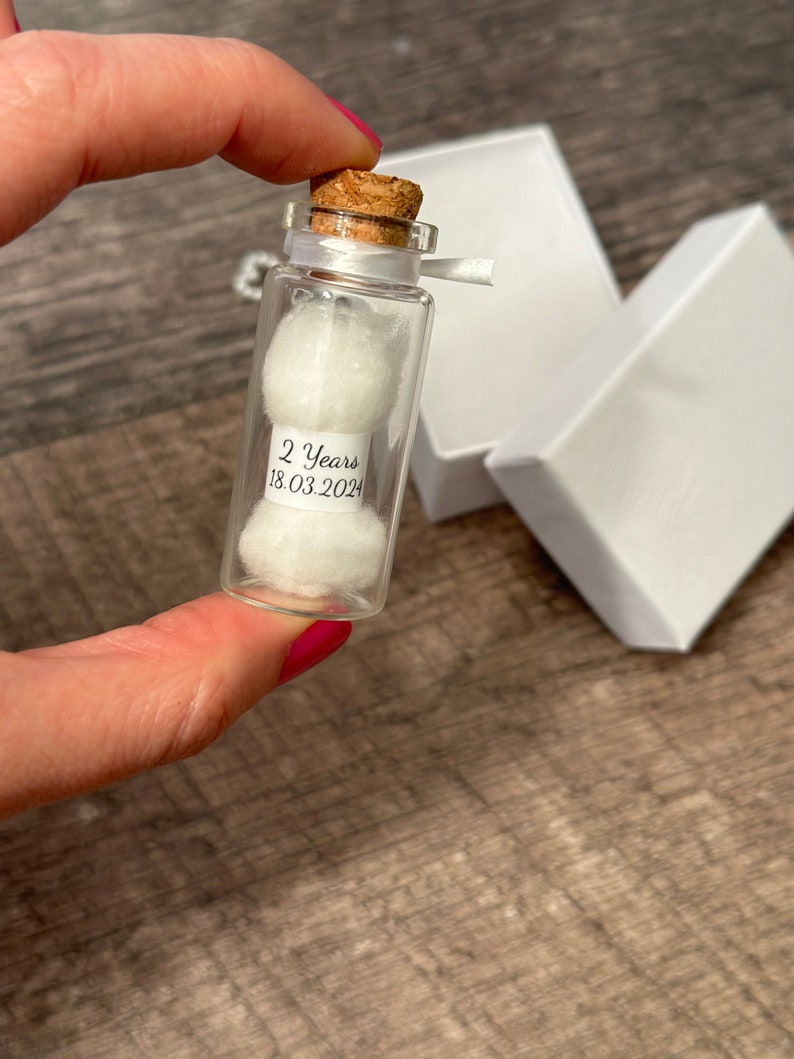 Op de afbeelding: Een klein glazen flesje met een kurkstop en een wit lint. Het flesje bevat twee witte wattenbolletjes en een label met de tekst "2 Years 18.03.2024".