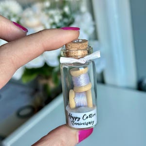 May include: A small glass bottle with a cork top filled with two spools of white thread. The bottle has a white label with the text "Happy Cotton Anniversary".