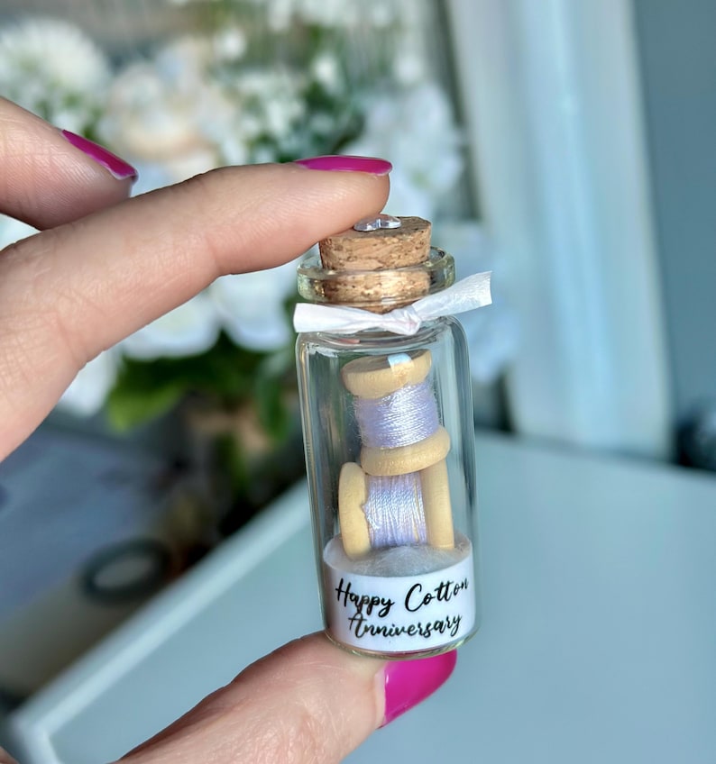 May include: A small glass bottle with a cork top filled with two spools of white thread. The bottle has a white label with the text "Happy Cotton Anniversary".