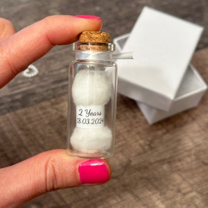 Op de afbeelding: Een klein glazen flesje met een kurkstop gevuld met witte wattenbolletjes. Een label op de fles staat "2 Years 18.03.2024".