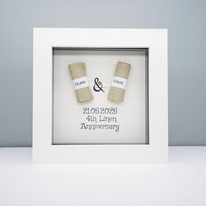 May include: A white framed artwork featuring two burlap rolls, one labelled "Lauren" and the other "David", with an ampersand between them. The text below reads "21.05.2025 4th Linen Anniversary". The background is a light beige.