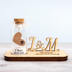 May include: A wooden anniversary gift. It includes a glass bottle with a cork, a heart-shaped tag, and the text "3 Years 08.03.2026". The wooden base has the initials "I & M" and the words "Happy Anniversary".