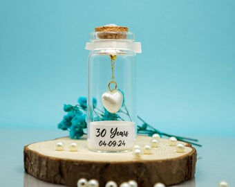 May include: A small glass bottle with a cork stopper contains a white heart charm on a gold chain. The bottle has a white label with the text "30 Years 04-09-24". The bottle is surrounded by white pearls on a wooden surface.