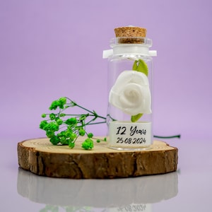 May include: A small glass bottle with a cork stopper contains a white fabric rose and a green leaf. The bottle has a white label with the text "12 Years 25-08-2024".