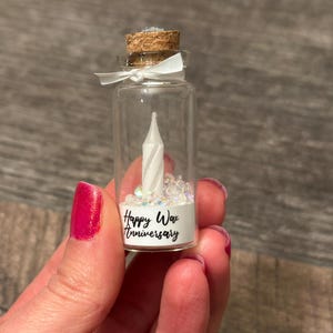 May include: A small glass bottle with a cork top filled with white glitter and a white wax candle. The bottle has a label that reads "Happy Wax Anniversary".