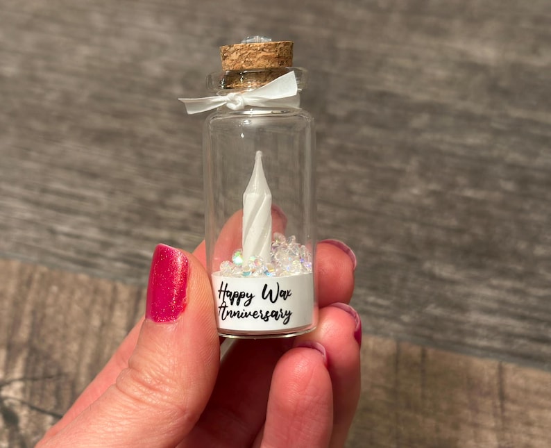 May include: A small glass bottle with a cork top filled with white glitter and a white wax candle. The bottle has a label that reads "Happy Wax Anniversary".