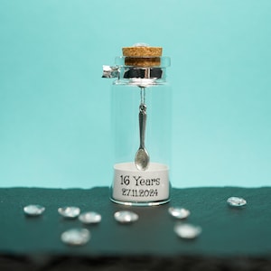 May include: A small glass jar with a cork top and a silver spoon hanging inside. The jar has a white label with the text "16 Years 27.11.2024" printed on it.