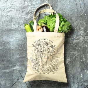 May include: A beige canvas tote bag filled with fresh produce, including zucchini, broccoli, and leafy greens. The bag features a black illustration of three women and the text "SASSY GIRLS CLUB". The bag has a handle for carrying.