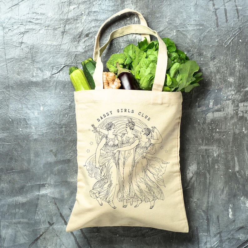 May include: A beige canvas tote bag filled with fresh produce, including zucchini, broccoli, and leafy greens. The bag features a black illustration of three women and the text "SASSY GIRLS CLUB". The bag has a handle for carrying.