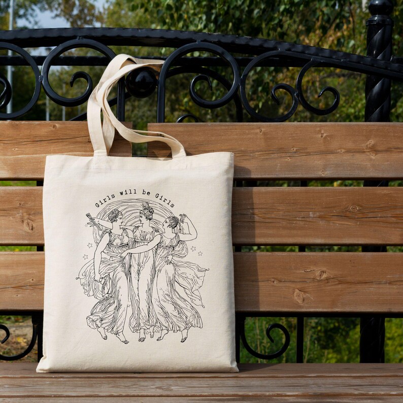 May include: A cream-colored tote bag with the text "Girls will be Girls" above a black and white illustration of three women in flowing gowns. The bag has a rectangular shape and two handles.
