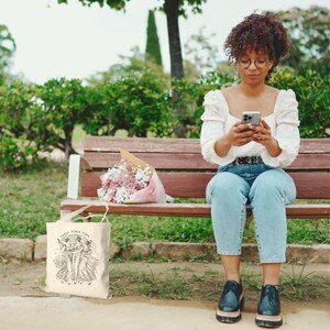May include: A cream-colored tote bag with the text "Saint Girls Club" and a graphic of two figures in flowing dresses. The bag is sitting on the ground next to a wooden bench. A bouquet of flowers rests on the bench.