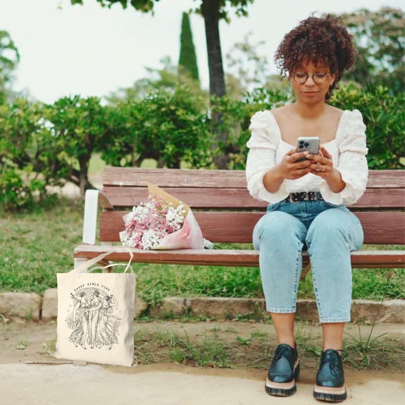 May include: A cream-colored tote bag with the text "Saint Girls Club" and a graphic of two figures in flowing dresses. The bag is sitting on the ground next to a wooden bench. A bouquet of flowers rests on the bench.