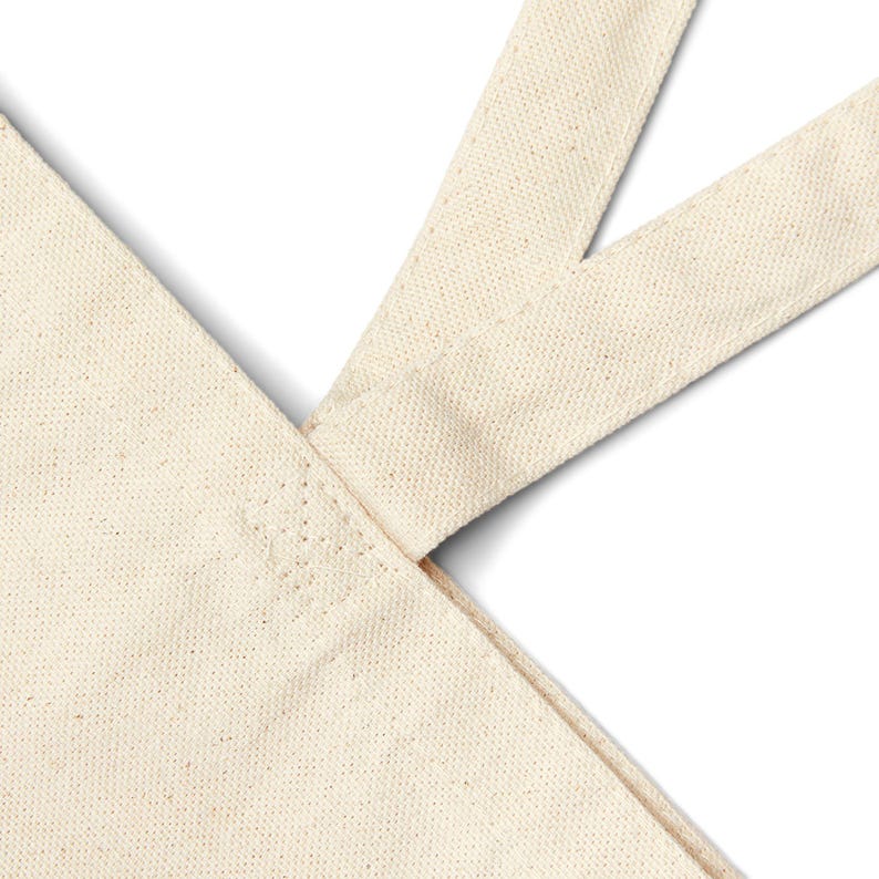 May include: Close-up of a natural canvas tote bag. The bag features a cream-colored fabric with a textured weave. The straps are attached at the top corners, creating a V-shape. The stitching is visible along the seams.