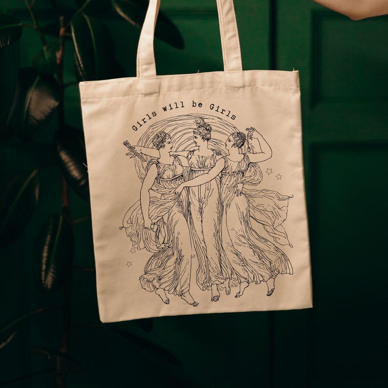 May include: Beige tote bag with the text "Girls will be Girls" above a black and white illustration of three women in flowing gowns. The women are in a circle, with stars around them.