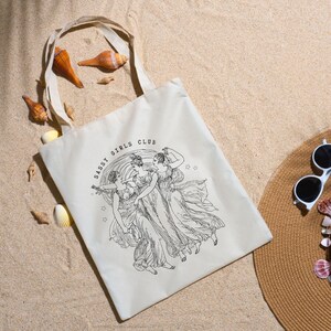 May include: Cream-colored tote bag with the text "Sassy Girls Club" and a black and white illustration of three women in flowing gowns. The bag is on a sandy surface with seashells and a straw hat.