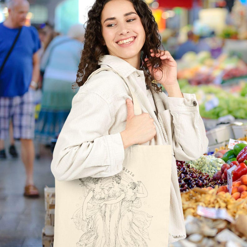 May include: A beige tote bag with a black and white illustration of three figures and the word "girls" printed above them. The bag is carried by a person in a light-colored jacket, smiling in an outdoor market setting.