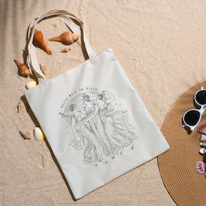 May include: Off-white tote bag with a black illustration of three women in flowing gowns, with the text "Girls will be Girls". The bag has a natural canvas texture and a simple handle. Seashells and a sun hat are in the background.