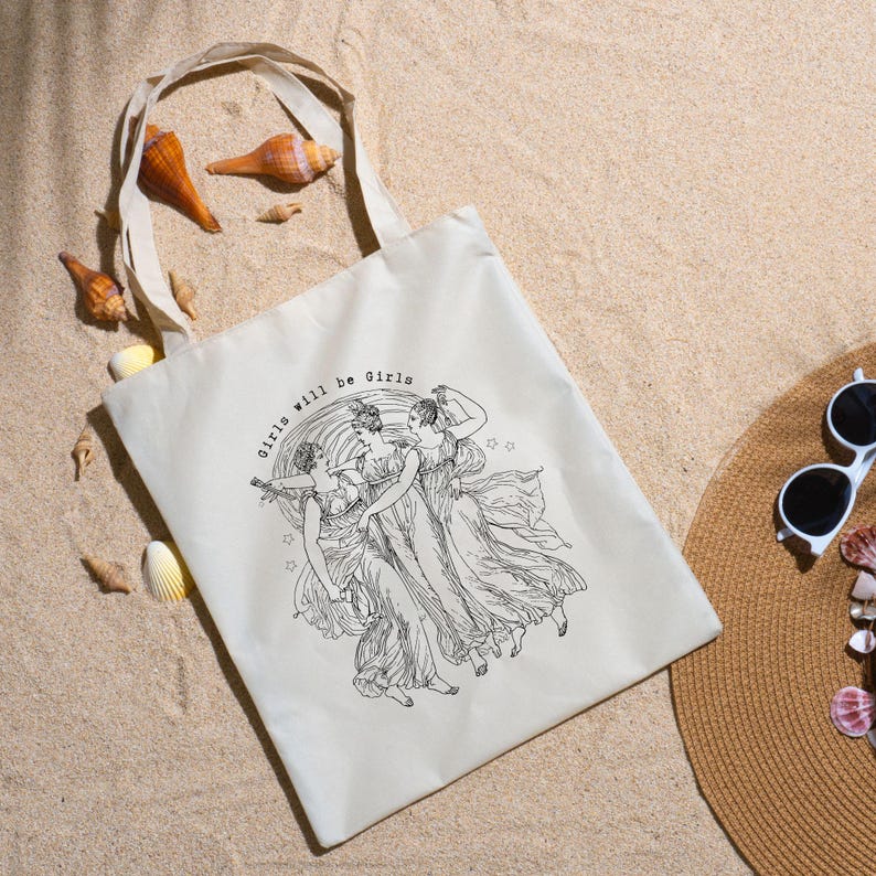 May include: Off-white tote bag with a black illustration of three women in flowing gowns, with the text "Girls will be Girls". The bag has a natural canvas texture and a simple handle. Seashells and a sun hat are in the background.