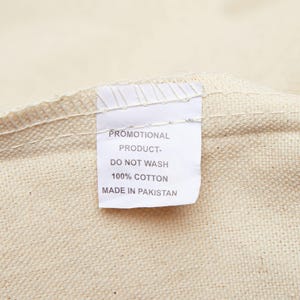May include: Close-up of a cream-colored fabric with a white tag. The tag reads "PROMOTIONAL PRODUCT - DO NOT WASH - 100% COTTON - MADE IN PAKISTAN". The fabric has a textured appearance.