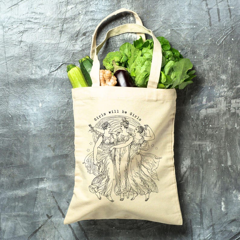 May include: A beige canvas tote bag filled with fresh produce, including zucchini, broccoli, and leafy greens. The bag features a black and white illustration of three women and the text "Girls will be Girls".