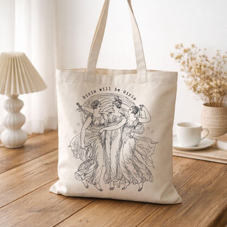 May include: Cream-colored tote bag featuring a black illustration of three women in flowing gowns, with the text "Girls will be Girls" above the image. The bag has two handles for carrying.