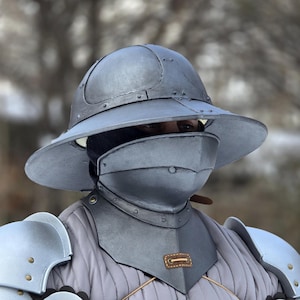 May include: A full suit of medieval armor in silver and gray, including a helmet with a wide brim, face guard, and shoulder, arm, and chest protection. The armor is detailed with rivets, leather straps, and gold accents. The undergarment is a quilted gray fabric.